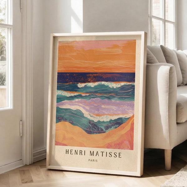 May include: A framed art print leaning against a beige couch in a bright living room with white walls and windows. The colorful poster shows an abstract seascape with orange sunset sky, dark blue horizon, teal ocean waves with white foam, and sandy beach in warm tones. Text reads "HENRI MATISSE PARIS" at bottom. Light wood frame, approximately 2-3 feet tall.