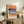 May include: Colorful abstract seascape poster with orange sunset sky, blue horizon, turquoise waves with white foam, and sandy beach in warm tones. Framed print displayed on rustic wood sideboard alongside fern plant, ceramic vase, books, and cream lamp in cozy living room setting with neutral walls and leather furniture.