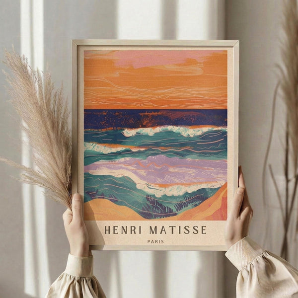 May include: Person in beige clothing holding framed art print featuring abstract seascape with orange sunset sky, teal rolling waves, and sandy beach in layered collage style. Natural wood frame against white curtained window with dried pampas grass decoration visible to left.