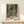 May include: Framed vintage-style art print featuring tropical foliage viewed through open French doors, displaying vibrant greens, oranges, and reds with palm fronds and exotic plants. Print shows "HENRI MATISSE" text at bottom, displayed in thin wooden frame on light wood surface beside small white wrapped gift with evergreen sprig against cream wall.