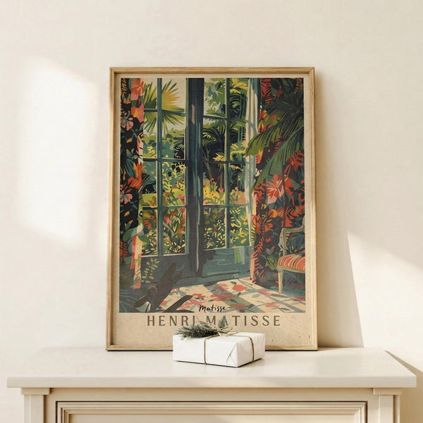 May include: Framed vintage-style art print featuring tropical foliage viewed through open French doors, displaying vibrant greens, oranges, and reds with palm fronds and exotic plants. Print shows "HENRI MATISSE" text at bottom, displayed in thin wooden frame on light wood surface beside small white wrapped gift with evergreen sprig against cream wall.