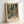 May include: Framed art print in light wood frame leaning against white wall. Artwork shows tropical indoor scene with French doors opening to lush garden filled with colorful palm fronds, exotic plants and flowers in vibrant greens, oranges and reds. Patterned tile floor and furniture visible. Text reads "HENRI MATISSE PARIS" at bottom in vintage poster style.