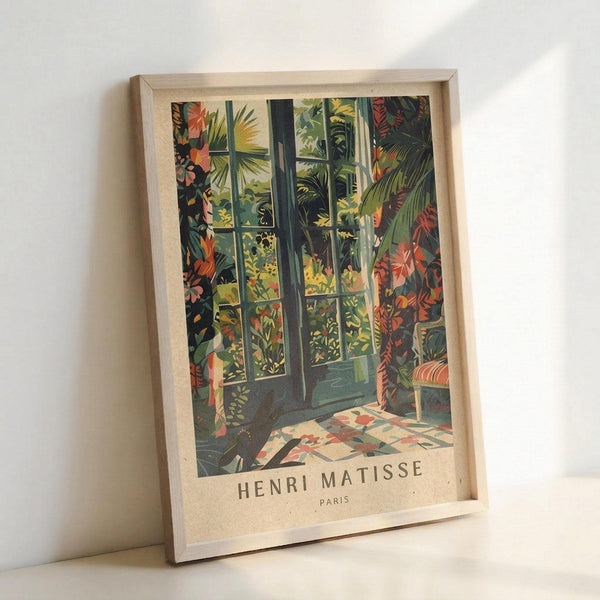 May include: Framed art print in light wood frame leaning against white wall. Artwork shows tropical indoor scene with French doors opening to lush garden filled with colorful palm fronds, exotic plants and flowers in vibrant greens, oranges and reds. Patterned tile floor and furniture visible. Text reads "HENRI MATISSE PARIS" at bottom in vintage poster style.