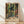 May include: Framed art print in light wood frame leaning against white wall on light wooden floor. Print shows tropical indoor garden scene with large windows, lush green palm fronds, colorful foliage in oranges and reds, and geometric patterned floor tiles. Warm sunlight streams through glass doors revealing dense jungle vegetation outside. Henri Matisse Paris text appears at bottom of vintage-style poster.
