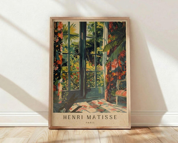 May include: Framed art print in light wood frame leaning against white wall on light wooden floor. Print shows tropical indoor garden scene with large windows, lush green palm fronds, colorful foliage in oranges and reds, and geometric patterned floor tiles. Warm sunlight streams through glass doors revealing dense jungle vegetation outside. Henri Matisse Paris text appears at bottom of vintage-style poster.