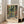May include: Framed poster print featuring tropical garden scene with lush green foliage, orange and red flowers, and French doors opening to verdant landscape. Print displays "Henri Matisse Paris" text at bottom with vintage beige border. Light wood frame leans against white wall next to beige sofa in bright, modern living room with large windows.