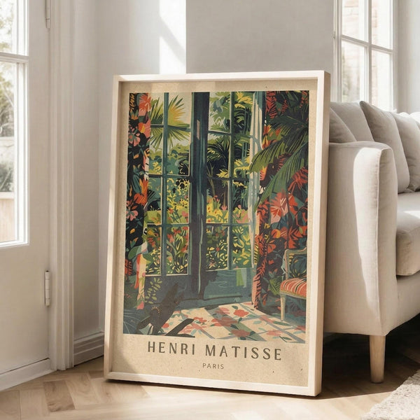 May include: Framed poster print featuring tropical garden scene with lush green foliage, orange and red flowers, and French doors opening to verdant landscape. Print displays "Henri Matisse Paris" text at bottom with vintage beige border. Light wood frame leans against white wall next to beige sofa in bright, modern living room with large windows.
