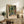 May include: Framed Henri Matisse Paris poster displaying vibrant tropical foliage and French doors in greens, oranges, and yellows, positioned on rustic wooden sideboard alongside fern plant, ceramic vase, stacked books, and beige lamp in warmly lit living room with cream chair and throw pillows