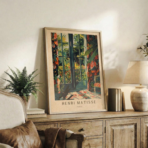 May include: Framed Henri Matisse Paris poster displaying vibrant tropical foliage and French doors in greens, oranges, and yellows, positioned on rustic wooden sideboard alongside fern plant, ceramic vase, stacked books, and beige lamp in warmly lit living room with cream chair and throw pillows