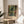 May include: Framed Henri Matisse art print displayed on round wooden side table next to beige upholstered chair. Poster features vibrant tropical scene with lush green palms, orange and red flowers viewed through dark window frames. Natural sunlight casts geometric shadows across cream walls. Plant leaves visible in foreground create layered composition in minimalist modern interior setting.