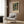May include: Framed Henri Matisse poster print featuring tropical foliage and interior scene with green palm fronds and colorful plants, displayed on wood-paneled wall above beige upholstered armchair with geometric patterned pillow, alongside brass floor lamp and dark round side table with open book