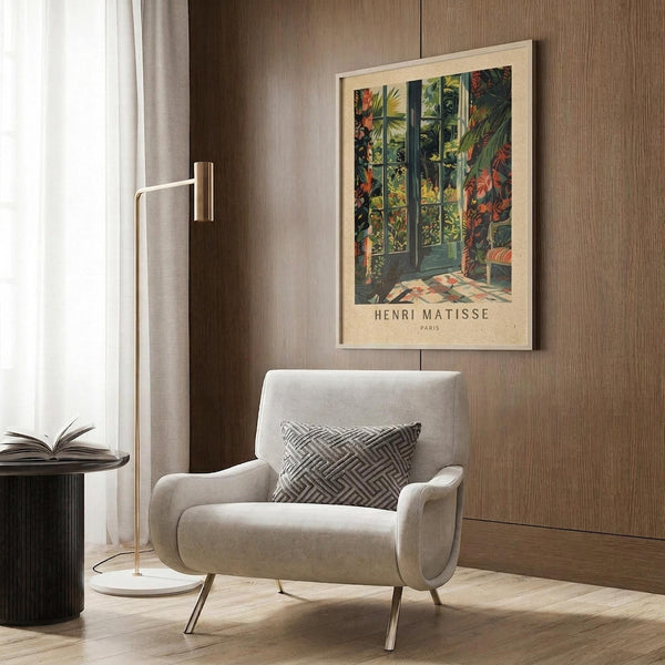 May include: Framed Henri Matisse poster print featuring tropical foliage and interior scene with green palm fronds and colorful plants, displayed on wood-paneled wall above beige upholstered armchair with geometric patterned pillow, alongside brass floor lamp and dark round side table with open book