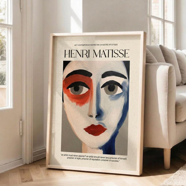 May include: Large framed art print leaning against white wall next to beige sofa in bright living room. Print shows stylized portrait with red and blue color blocks around eyes, red lips, and Henri Matisse text at top. Light wood frame on hardwood floors with natural window lighting.