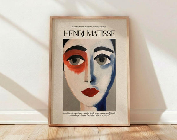 May include: Framed poster featuring a stylized portrait in Matisse-inspired style with bold colors - red and blue face tones, black eyebrows, red lips, against cream background. Text reads "HENRI MATISSE" at top with French subtitle and quote below. Natural wood frame sits on light wooden floor against white wall with soft window light shadows.