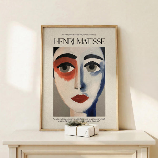 May include: Framed art print featuring an abstract portrait with asymmetrical face in red, blue and white tones, displayed on light wooden console table beside small wrapped gift with twine bow, against neutral beige wall with natural lighting