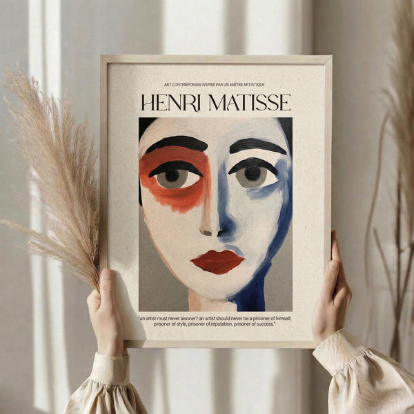 May include: Person in cream ruffled blouse holding framed art print featuring stylized portrait with asymmetrical face design in red, blue, and neutral tones. Modern minimalist frame against soft curtained background. Person also holds dried pampas grass stems. Contemporary art poster with French text and artist quote below the portrait illustration.
