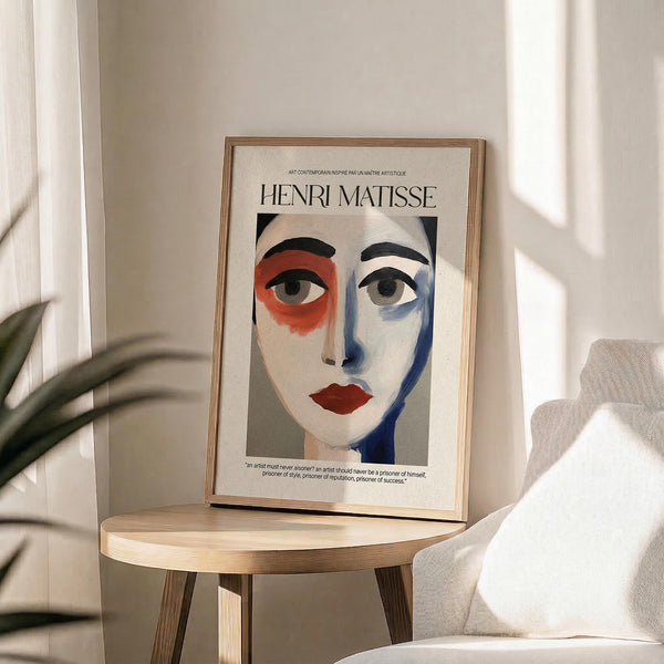 May include: Framed Henri Matisse-style portrait print displayed on light wood round side table. The artwork features an abstract face with asymmetrical coloring - orange-red around one eye, blue shadowing on the other side, red lips, and black eyebrows against cream background. Natural lighting creates soft shadows on beige wall behind. Green plant leaves visible in corner, neutral fabric chair arm on right.