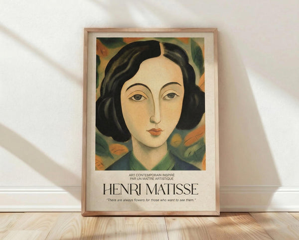 May include: Framed art print featuring a portrait of a woman with black bob haircut and serene expression, painted in warm earth tones with colorful floral background in greens and oranges. The poster displays "HENRI MATISSE" text and French description below the artwork. Natural wood frame sits on light wooden floor against white textured wall with soft natural lighting creating gentle shadows.