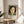 May include: Framed art print featuring a portrait of a woman with black bob haircut and stylized facial features in warm earth tones and greens, displayed on a round wooden side table next to a beige upholstered chair. Natural sunlight streams across the neutral-toned room setting with white curtains and a plant visible in the corner.
