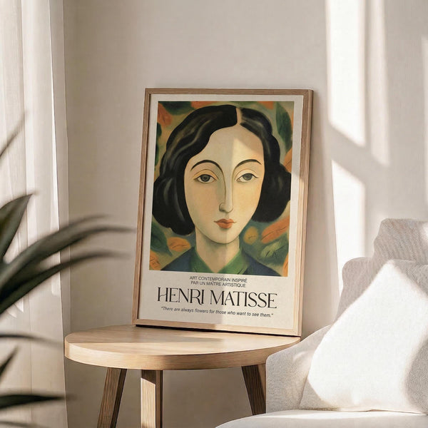 May include: Framed art print featuring a portrait of a woman with black bob haircut and stylized facial features in warm earth tones and greens, displayed on a round wooden side table next to a beige upholstered chair. Natural sunlight streams across the neutral-toned room setting with white curtains and a plant visible in the corner.