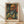 May include: Framed art print leaning against white wall on light wood floor. Features a colorful expressionist self-portrait of a bald man with mustache and beard, painted in bold orange, green, and blue tones. The artwork is displayed in a natural wood frame and shows geometric, angular brushwork typical of early modern art style.
