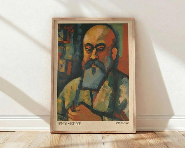 May include: Framed art print leaning against white wall on light wood floor. Features a colorful expressionist self-portrait of a bald man with mustache and beard, painted in bold orange, green, and blue tones. The artwork is displayed in a natural wood frame and shows geometric, angular brushwork typical of early modern art style.