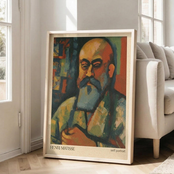 May include: Framed art print of a colorful self-portrait showing a bald man with mustache and beard in Post-Impressionist style, featuring vibrant oranges, greens, and blues. The wooden-framed poster leans against a white wall next to a beige sofa in a bright, minimalist living room with large windows and natural lighting.