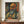 May include: Framed art print displaying a colorful expressionist-style self portrait of a bald man with mustache and beard, rendered in vibrant oranges, greens, and blues. The poster sits on a wooden sideboard next to a white ceramic vase and books, against cream wainscoted walls in an elegant interior setting.