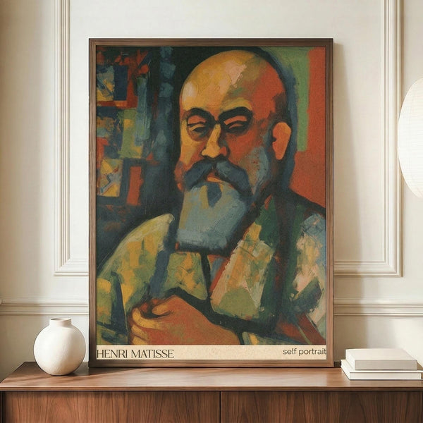 May include: Framed art print displaying a colorful expressionist-style self portrait of a bald man with mustache and beard, rendered in vibrant oranges, greens, and blues. The poster sits on a wooden sideboard next to a white ceramic vase and books, against cream wainscoted walls in an elegant interior setting.