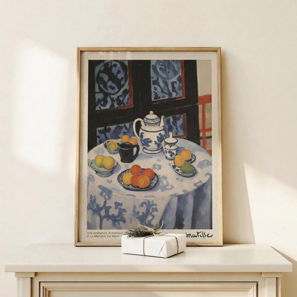 May include: Framed Henri Matisse still life print displayed on light wood console table against cream wall. Artwork shows round table with white patterned tablecloth, blue and white teapot set, colorful fruit on plates, and decorative tiles on dark wall behind. Natural wood frame with white matting. Small wrapped gift box with greenery sits in front of frame.