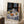 May include: Framed Henri Matisse still life print leaning against white wall in cozy living room setting. The artwork shows a round table with blue patterned tablecloth, featuring oranges, teapot, cups, and decorative wall tiles in background. Natural wood frame complements the blue, white, and orange color palette. White coffee cup sits on wooden side table nearby, with soft beige textiles visible.