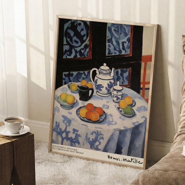 May include: Framed Henri Matisse still life print leaning against white wall in cozy living room setting. The artwork shows a round table with blue patterned tablecloth, featuring oranges, teapot, cups, and decorative wall tiles in background. Natural wood frame complements the blue, white, and orange color palette. White coffee cup sits on wooden side table nearby, with soft beige textiles visible.