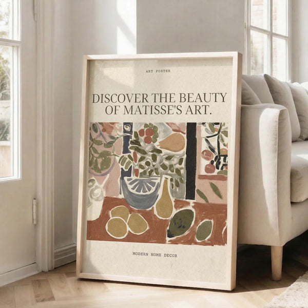 May include: Framed art poster featuring a colorful Matisse-style still life with plants, fruit, and geometric shapes in earthy tones of green, terracotta, and cream. The poster has text reading "Discover the Beauty of Matisse's Art" and "Modern Home Decor." Light wood frame leans against white wall next to beige sofa in bright, airy room with large windows and natural sunlight streaming across light wooden floors.