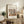 May include: Framed Matisse-style art poster displayed on rustic wooden sideboard in neutral living room. Poster features colorful still life with botanical elements, vase, and fruit in earthy tones. Scene includes fern plant, ceramic vases, stacked books, and brown leather furniture. Natural lighting creates warm, cozy atmosphere with beige walls.