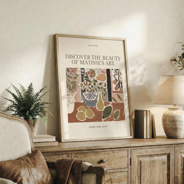 May include: Framed Matisse-style art poster displayed on rustic wooden sideboard in neutral living room. Poster features colorful still life with botanical elements, vase, and fruit in earthy tones. Scene includes fern plant, ceramic vases, stacked books, and brown leather furniture. Natural lighting creates warm, cozy atmosphere with beige walls.
