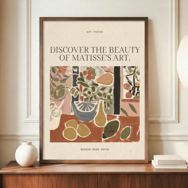 May include: Framed art poster featuring a Matisse-style still life with plants in a decorative vase, fruits, and tabletop objects in earthy tones of terracotta, olive green, and cream. The poster has text reading "Discover the Beauty of Matisse's Art" and "Modern Home Decor." Displayed on a wooden sideboard with paneled walls, alongside a small cream ceramic vase and stacked books.