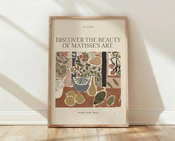 May include: Framed art poster with cream background featuring colorful Matisse-style still life artwork showing fruits, plants, and vessels in earthy tones of green, brown, and terracotta. Text reads "Discover the beauty of Matisse's art" and "Modern home decor." Natural wood frame sits on light wooden floor against white wall with soft window light creating shadows.