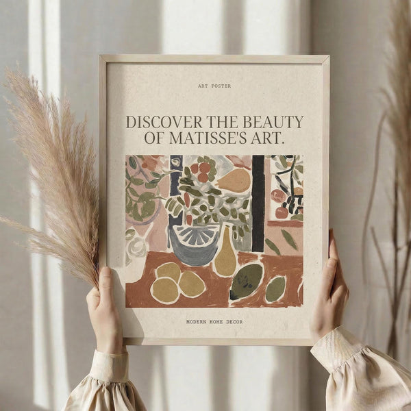 May include: Hands holding a framed art poster featuring Matisse-inspired still life with botanical elements, fruits, and vessels in muted earth tones of terracotta, sage green, and cream. The poster has text about discovering beauty in art and modern home decor. Dried pampas grass stems visible on left side against soft neutral curtained background.