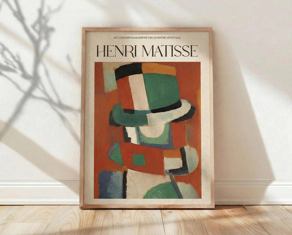 May include: Framed Henri Matisse poster print leaning against white wall on wooden floor. Abstract artwork features geometric shapes in vibrant colors - orange, green, red, blue, and white forms suggesting a figure wearing a hat. Natural wood frame with cream matting. Soft plant shadows cast on wall from window light.
