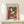 May include: Framed Henri Matisse poster featuring an abstract figure wearing a colorful hat in green, orange, red, and black brushstrokes against an orange background. The artwork is displayed in a light wood frame on a cream-colored shelf next to a small white wrapped gift, positioned against a white wall in minimalist home decor setting.