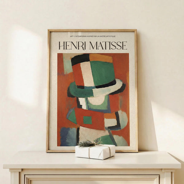 May include: Framed Henri Matisse poster featuring an abstract figure wearing a colorful hat in green, orange, red, and black brushstrokes against an orange background. The artwork is displayed in a light wood frame on a cream-colored shelf next to a small white wrapped gift, positioned against a white wall in minimalist home decor setting.