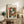May include: Framed Henri Matisse poster featuring abstract colorful composition with green, orange, red and blue geometric shapes suggesting a figure. Artwork displayed on rustic wooden sideboard with drawers alongside potted fern plant, ceramic vase, and stacked books. Neutral cream colored room setting with beige upholstered chair visible in foreground. Natural lighting creates soft shadows on white wall behind the framed print.