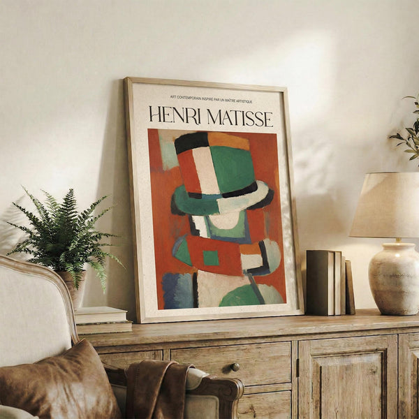 May include: Framed Henri Matisse poster featuring abstract colorful composition with green, orange, red and blue geometric shapes suggesting a figure. Artwork displayed on rustic wooden sideboard with drawers alongside potted fern plant, ceramic vase, and stacked books. Neutral cream colored room setting with beige upholstered chair visible in foreground. Natural lighting creates soft shadows on white wall behind the framed print.