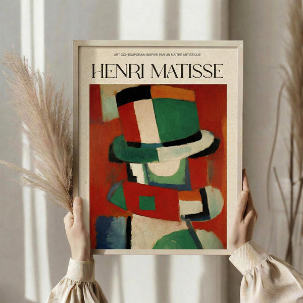 May include: Hands holding a framed art print featuring an abstract figure in a colorful hat, displayed in Matisse's distinctive style with bold red, green, white, and orange geometric shapes. The beige wooden frame is shown against flowing curtains with dried pampas grass visible to the left, creating a warm, natural interior setting.