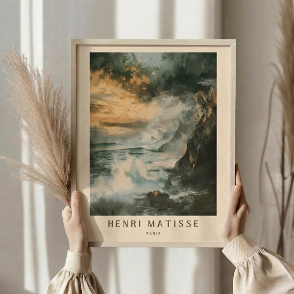 May include: Person holding framed art print showing dramatic coastal seascape with turbulent storm clouds in orange and green tones, rocky cliffs, and choppy ocean waves. Light wooden frame with cream matting displays "Henri Matisse Paris" text below romantic landscape artwork. Person wears cream ruffled sleeve top, holds dried pampas grass stems, standing by bright window with white curtains.
