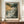 May include: Framed poster displaying a dramatic seascape with stormy clouds, rocky cliffs, and turbulent waves in muted greens, grays, and warm orange tones. Birds fly across the moody sky. Text reads "HENRI MATISSE PARIS" below the artwork. The brown-framed print sits on a wooden console beside a white ceramic vase and stacked books against cream walls with white molding.