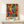 May include: Colorful abstract art poster in wooden frame showing dancing figures in vibrant blues, greens, oranges, and yellows with black silhouettes. The framed print displays "MATISSE" in green text below the artwork and sits on a light wood surface next to a small white wrapped gift with twine. Clean white wall background creates minimal modern display setting.
