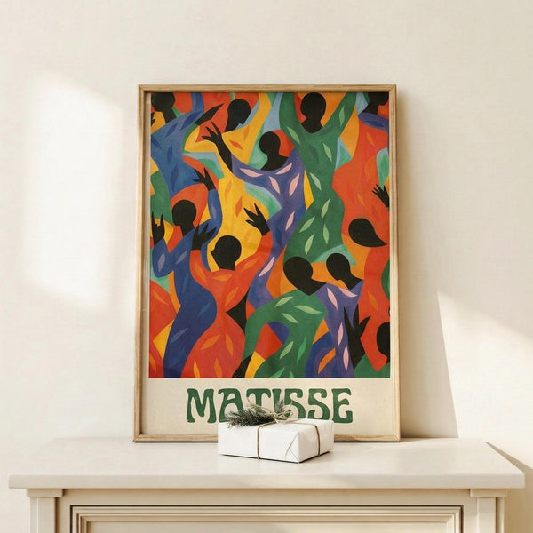 May include: Colorful abstract art poster in wooden frame showing dancing figures in vibrant blues, greens, oranges, and yellows with black silhouettes. The framed print displays "MATISSE" in green text below the artwork and sits on a light wood surface next to a small white wrapped gift with twine. Clean white wall background creates minimal modern display setting.