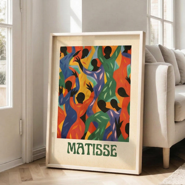 May include: Framed art print featuring vibrant abstract figures in flowing, dancing poses rendered in bold colors of orange, green, blue, purple and yellow with black silhouetted heads. The poster displays "MATISSE" in green text below the artwork and sits in a light wood frame against white walls near a beige upholstered sofa in a bright, naturally-lit room with large windows.
