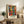 May include: Vibrant abstract art print featuring colorful dancing figures in blues, greens, oranges, and purples with black silhouetted heads, displayed in light wood frame on rustic wooden sideboard alongside fern plant, books, and ceramic vase in neutral-toned living room setting